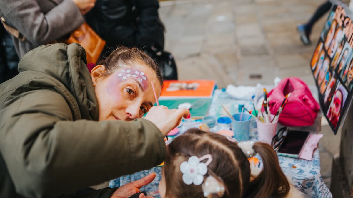 A face painter artistically decorates a child's face outdoors at a vibrant street festival.